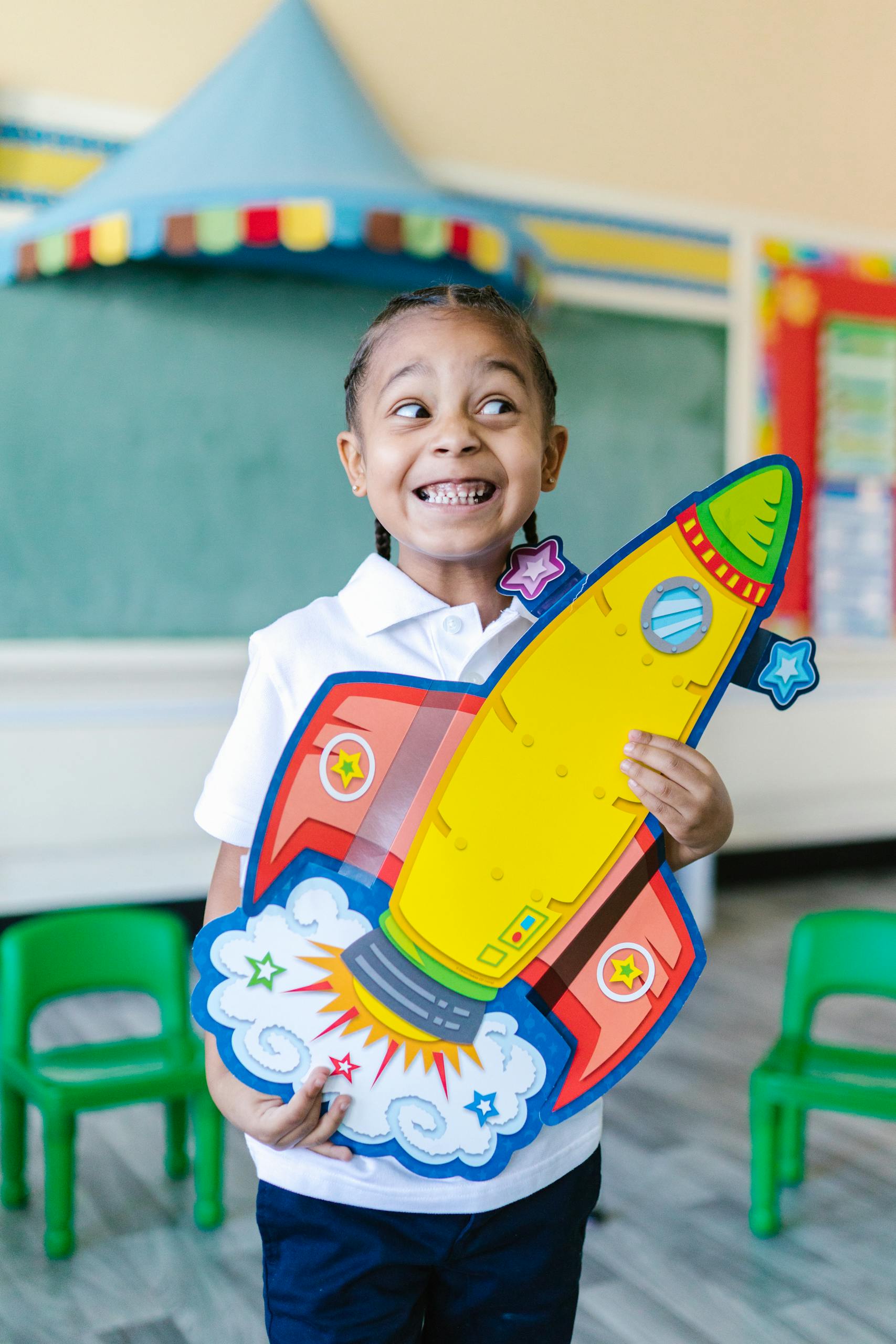 Smiling girl holding a rocket cutout in a vibrant classroom setting.