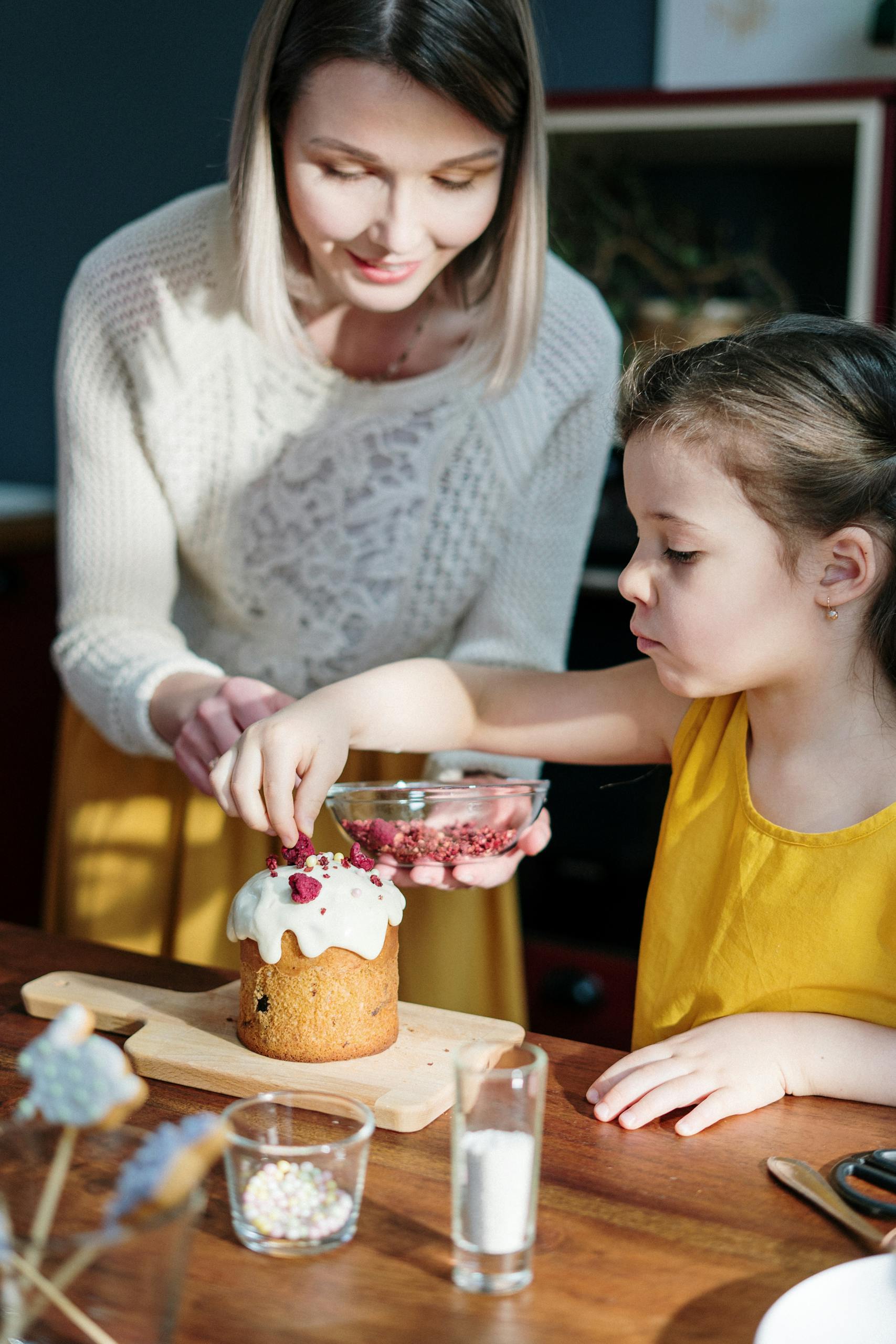 A mother and daughter enjoy decorating an Easter cake in a sunny kitchen.
